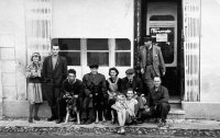 Family and friends in front of a house with a shop and glassworks in Vrbno pod Pradědem, Anna Marková sitting on the pavement with a little girl on her lap